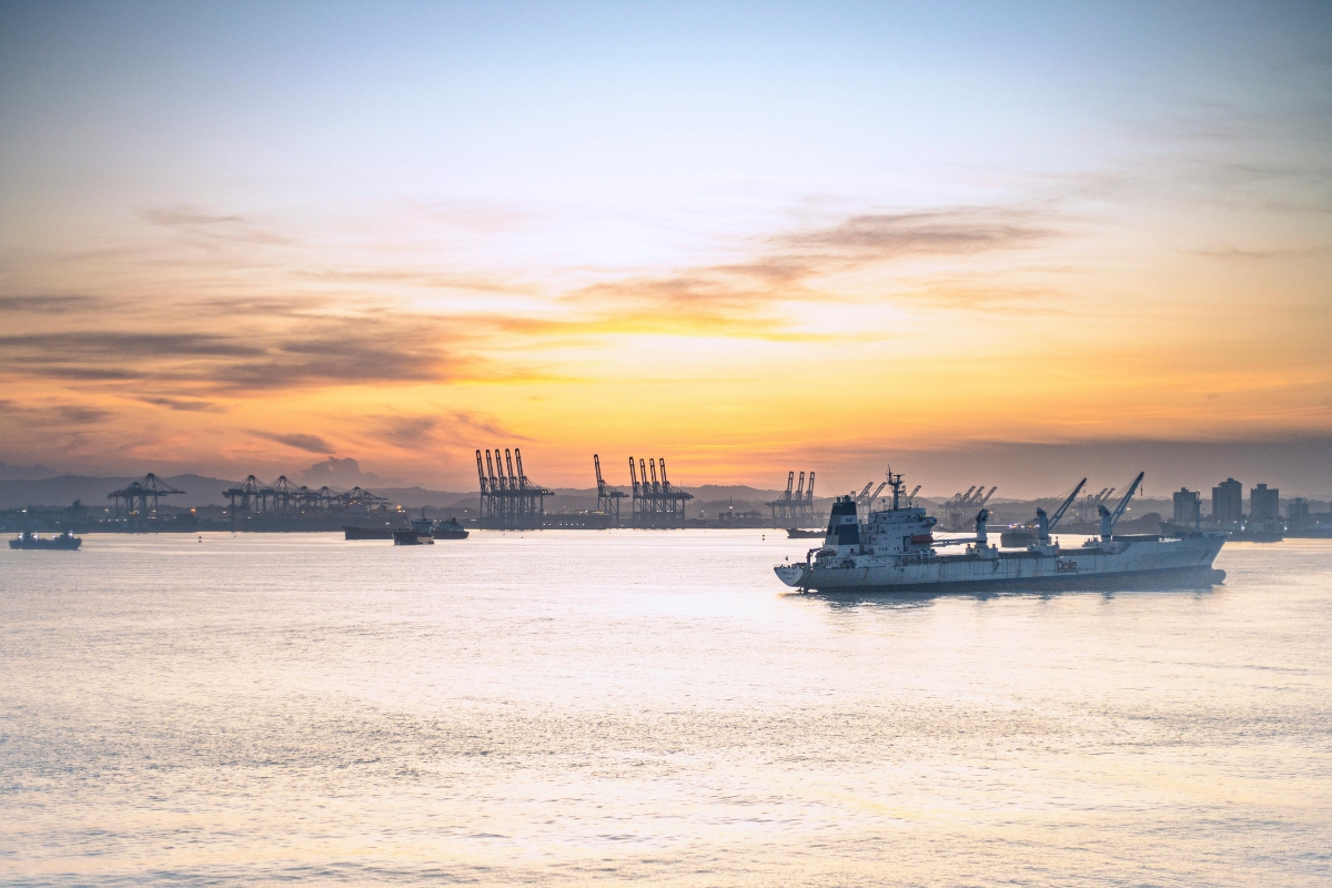Navio cargueiro atravessa águas calmas ao entardecer; guindastes do porto e cidade ao fundo.